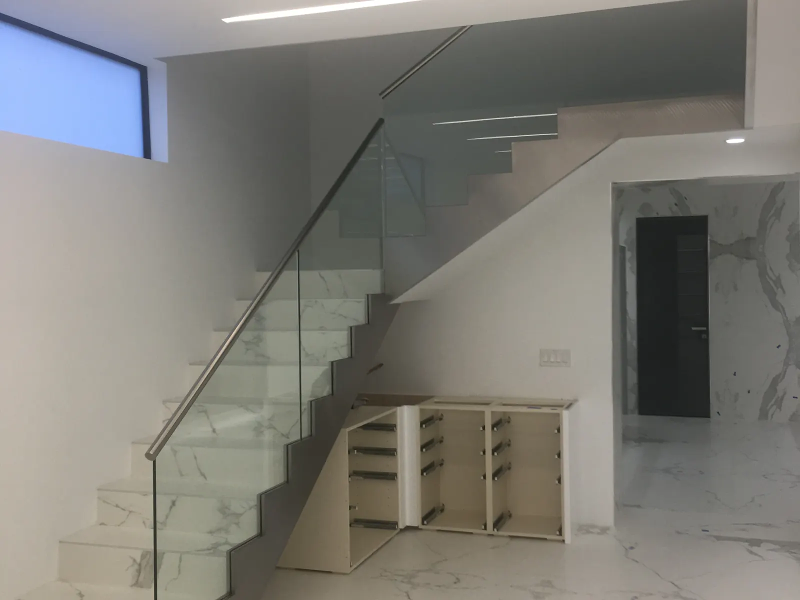 Interior remodel in progress with a marble-clad staircase, frameless glass railing and built-in cabinetry