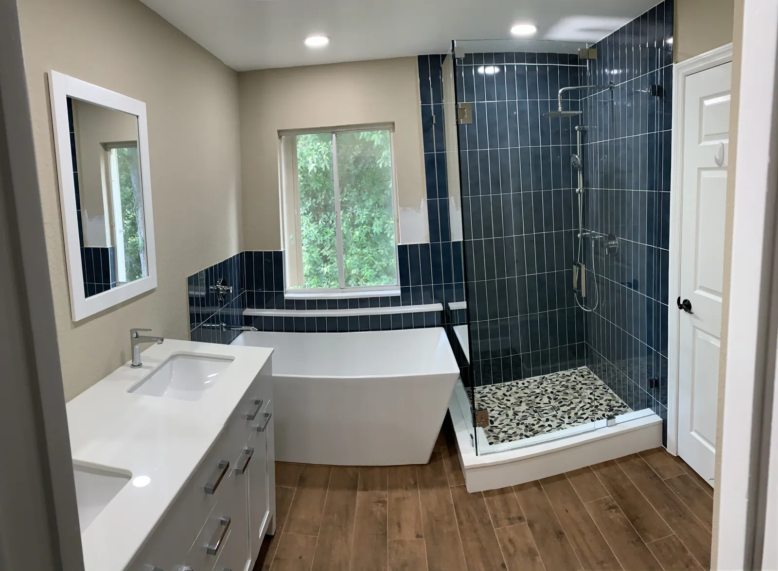Remodeled bathroom with a freestanding tub, frameless glass shower with navy wall tile and pebble shower floor, and wood-look floor tile